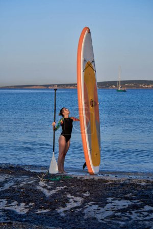 Téléchargez les photos : Vue de côté corps complet de surfeuse en maillot de bain portant la pagaie et debout avec SUP verticale sur le bord de la mer contre un paysage pittoresque - en image libre de droit