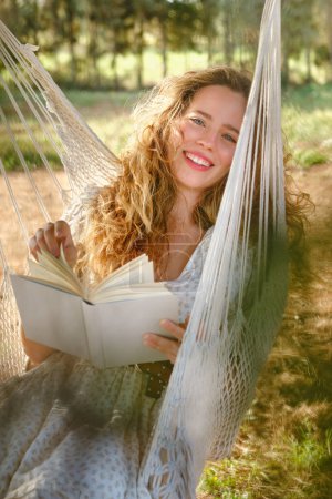 Téléchargez les photos : Optimiste jeune femme avec les cheveux longs souriant et regardant la caméra tout en lisant le bon livre sur l'hamac en week-end dans le jardin - en image libre de droit