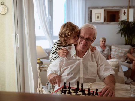 Téléchargez les photos : Garçon positif embrassant contenu grand-père assis à la table avec échiquier tout en jouant ensemble dans le salon léger à la maison - en image libre de droit