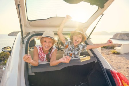 Téléchargez les photos : Deux enfants en chapeaux de paille assis sur les sièges arrière d'une voiture sortant la tête du coffre faisant des gestes de victoire avec les bras levés et une expression heureuse - en image libre de droit