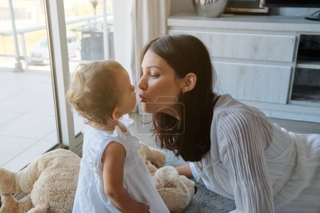 Photo for Side view of young mother in casual clothes sitting on floor and kissing cute little daughter while spending time at home - Royalty Free Image