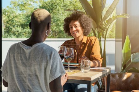 Téléchargez les photos : Amis féminines multiraciales positives en vêtements décontractés assis à la table en bois sur la terrasse du restaurant avec des verres de vin rouge et bavarder tout en se regardant et en attendant la nourriture - en image libre de droit
