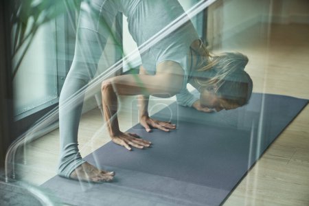 Téléchargez les photos : Grâce à un verre de femme en forme en vêtements de sport debout sur tapis gris et les jambes tendues dans Prasarita Padottasana Une pose tout en pratiquant le yoga dans la salle de gym - en image libre de droit