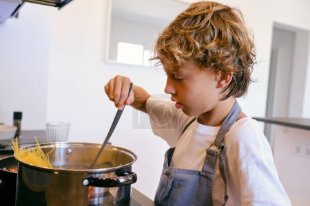 Photo for Side view of attentive child in apron with spatula cooking pasta in pot on stove in kitchen - Royalty Free Image