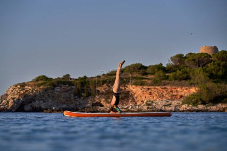 Téléchargez les photos : Vue latérale d'une athlète féminine sportive en maillot de bain prenant Salamba Shirshasana Une pose sur la planche SUP tout en pratiquant le yoga au milieu d'un océan bleu ondulant près d'une colline rocheuse au coucher du soleil - en image libre de droit
