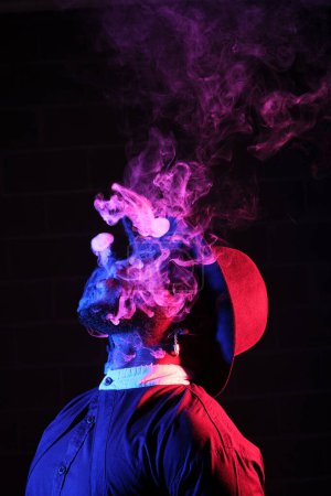 Foto de Vista lateral de un hombre irreconocible usando traje y peluca de pie y fumando en luz de neón - Imagen libre de derechos