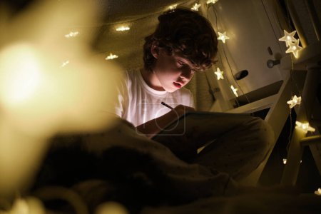 Photo for Focused talented boy drawing in sketchbook while sitting in blanket construction in dark room with glowing lamps at night time - Royalty Free Image