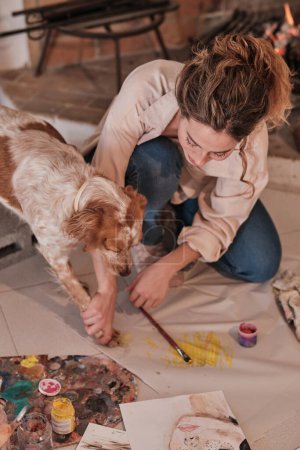 Foto de Artista femenina creativa vinculándose con mascotas usando pincel y estampado de patas mientras dibuja en tela en un estudio moderno - Imagen libre de derechos