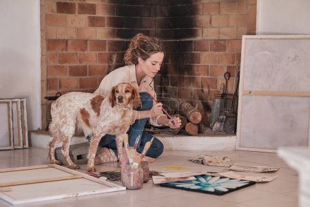 Photo for Side view of female artist with pet sitting on floor near fireplace and drawing using paintbrush and professional paints in creative workspace - Royalty Free Image