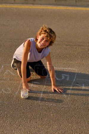 Téléchargez les photos : Délicieux préadolescent garçon dessin marelle avec de la craie sur la route tout en s'amusant en soirée d'été et en regardant la caméra - en image libre de droit