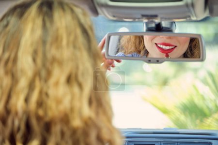 Téléchargez les photos : Retour joyeuse jeune conductrice appliquant rouge à lèvres rouge et réfléchissant dans le rétroviseur - en image libre de droit