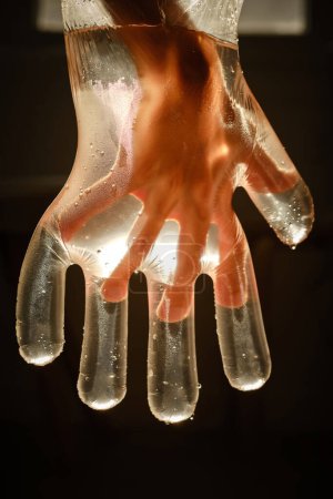Téléchargez les photos : Crop enfant anonyme dans un gant en plastique transparent avec de l'eau brillante et des gouttelettes sur fond sombre - en image libre de droit