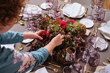 Téléchargez les photos : Angle élevé de culture Fleuriste féminine anonyme en chemisier arrangeant bouquet de fleurs fleuries assorties sur table de banquet avec verres et vaisselle - en image libre de droit