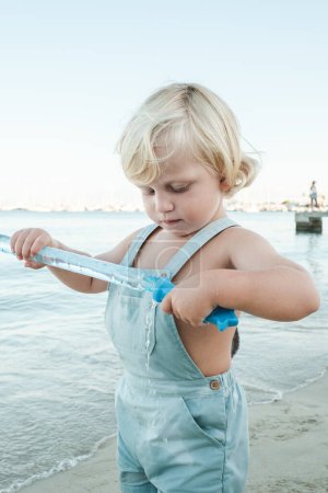 Téléchargez les photos : Adorable petit enfant explorant récipient avec bulles de savon sur le bord de la mer avec la côte de lavage de l'eau par temps ensoleillé - en image libre de droit