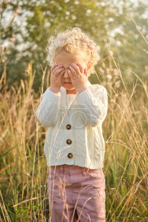 Photo for Cute little girl in warm sweater covering eyes with hands while standing on grassy lush lawn in sunny summer countryside - Royalty Free Image