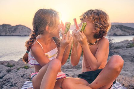 Foto de Feliz chico y chica en trajes de baño fingiendo sonreír con pieles de sandía y mirándose durante el picnic en la playa por la noche - Imagen libre de derechos