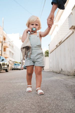 Photo for Full body of cute blond haired baby in overall and sandals with pacifier in mouth walking on asphalt road by hand with crop parent and holding toy - Royalty Free Image