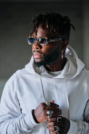 Photo for Serious black male model with dreadlocks wearing stylish hoodie and sunglasses looking away on blurred background - Royalty Free Image