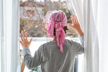 Téléchargez les photos : Vue arrière d'un enfant méconnaissable en bandana rose regardant la porte du balcon et touchant le verre à la lumière du soleil - en image libre de droit