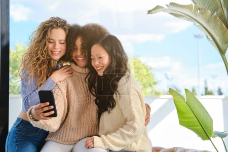 Photo for Content young diverse ladies in sweaters sitting close to each other and hugging while taking self portrait on mobile phone - Royalty Free Image