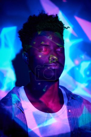 Téléchargez les photos : Homme afro-américain immobile portant une tenue décontractée debout avec les yeux fermés dans les lumières au néon dans le club sombre - en image libre de droit