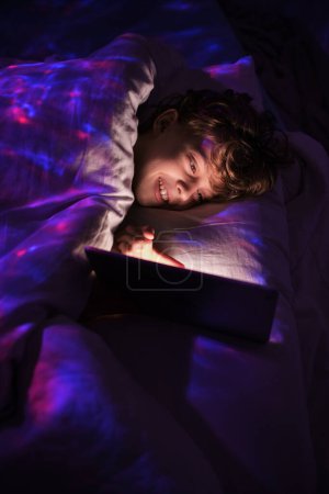 Photo for Content boy surfing tablet with smile while lying in bed under blanket in bedroom with dim light at night time - Royalty Free Image
