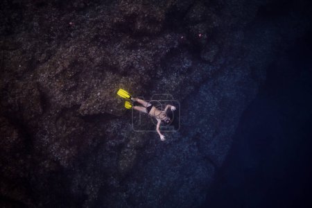 Téléchargez les photos : Vue de dessus corps complet de l'homme anonyme en masque de plongée avec tuba et palmes jaunes nageant sous l'eau de mer sombre profonde près de formation rocheuse massive - en image libre de droit