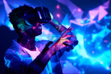 Foto de Glad hombre afroamericano vistiendo gafas VR y experimentando realidad virtual en luces de neón en el estudio - Imagen libre de derechos