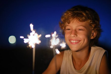Foto de Feliz niño preadolescente mirando las luces brillantes de Bengala mientras disfruta de la celebración de las vacaciones por la noche contra el cielo oscuro - Imagen libre de derechos