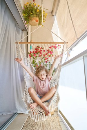 Foto de Cuerpo completo de adorable niño sentado en silla colgante en el balcón con los ojos cerrados y las piernas cruzadas y levantando brazos - Imagen libre de derechos