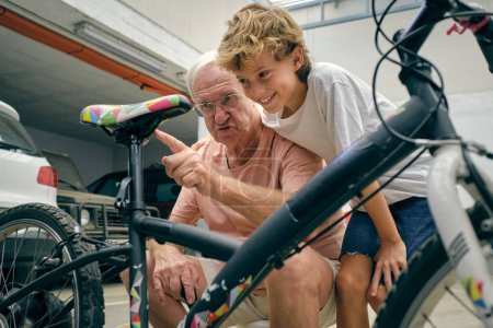 Téléchargez les photos : Grand-père sérieux expliquant comment réparer vélo à garçon écoute attentivement et souriant largement sur le parking - en image libre de droit