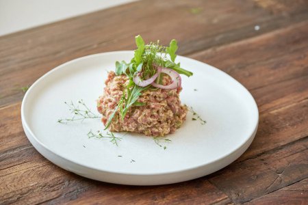 Photo for From above of appetizing steak tartar served with red onion and green herbs on white plate on wooden table - Royalty Free Image