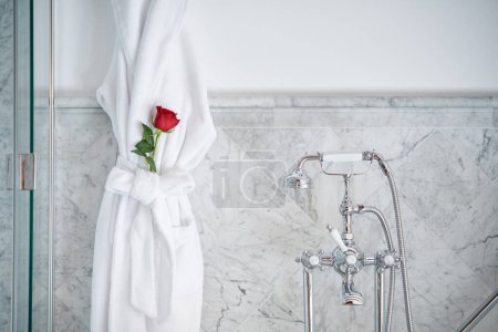 Téléchargez les photos : Intérieur de la salle de bain de luxe avec robinet vintage et peignoir en éponge blanc avec rose rouge placé à la ceinture suspendue sur des murs en marbre carrelé - en image libre de droit