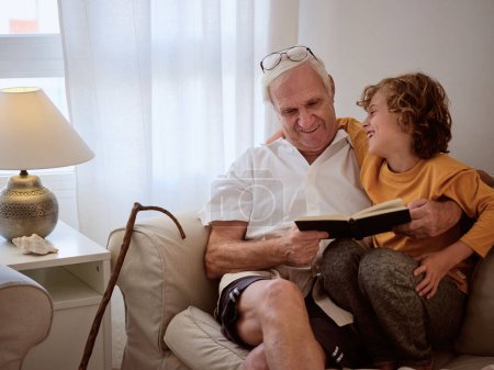 Foto de Feliz niño preadolescente abrazando contenido abuelo mientras descansan en cómodo sofá y libro de lectura juntos - Imagen libre de derechos