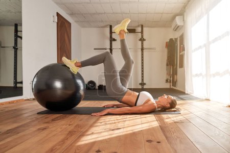 Téléchargez les photos : Vue latérale de la femme sportive en tenue de sport couchée sur le tapis et faisant des poussées de hanche avec la jambe levée sur la balle en forme pendant l'entraînement dans la salle de gym - en image libre de droit