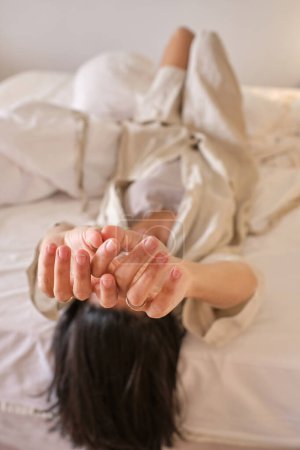 Téléchargez les photos : Mise au point douce de la femme méconnaissable en vêtements de nuit et peignoir couché sur le lit à l'envers et étirant les bras après le réveil le matin - en image libre de droit