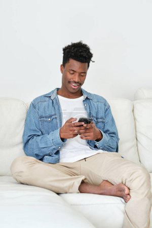 Foto de Joven hombre afroamericano alegre en ropa casual usando el teléfono móvil mientras se relaja en el cómodo sofá en la sala de estar de luz - Imagen libre de derechos