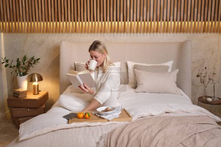 Photo for Full body of adult female in white sleepwear sitting on bed with tray and reading interesting book while drinking coffee in bedroom at home - Royalty Free Image