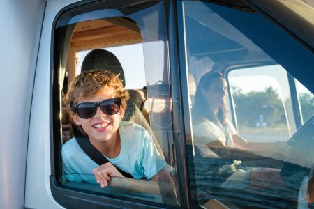 Photo for Smiling preteen boy in sunglasses peeking out of opened window of camper van and looking at camera while traveling with mother during summer vacation - Royalty Free Image