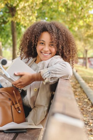 Téléchargez les photos : Joyeux jeune femme afro-américaine avec coiffure afro assis sur un banc en bois dans le parc d'automne avec un livre et regardant la caméra - en image libre de droit