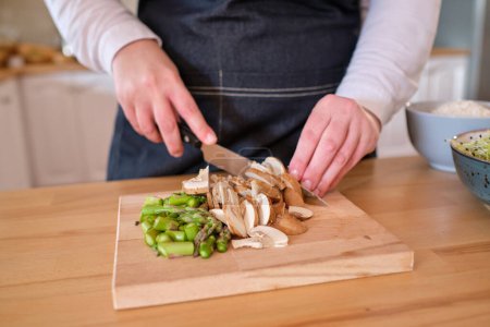 Photo for Crop cook in apron and with knife cutting raw mushrooms and asparagus on wooden chopping board while preparing vegetarian lunch in kitchen - Royalty Free Image