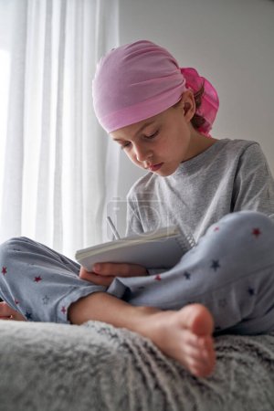 Photo for Serious boy in pajama and pink headscarf symbol of breast cancer awareness sitting with crossed legs and writing in notebook on sofa - Royalty Free Image