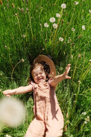 Foto de Desde arriba de la niña juguetona con sombrero de paja y en general acostado en prado herboso con dientes de león blancos en el día de verano soleado - Imagen libre de derechos