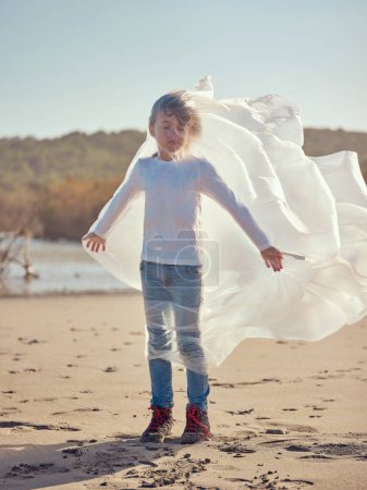 Foto de Cuerpo completo de niño rubio tranquilo con los ojos cerrados y la bolsa de plástico revoloteando alrededor del cuerpo de pie en la orilla de arena en el día soleado - Imagen libre de derechos