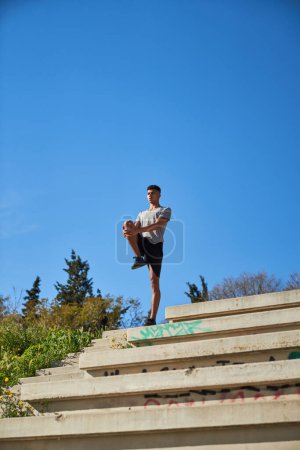 Photo for Distant sportsman doing warm up exercise with raised leg while standing on staircase against blue sky during training in city - Royalty Free Image