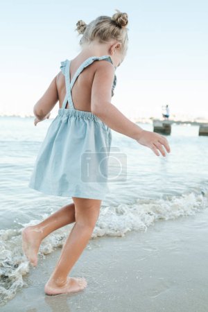 Photo for Side view of anonymous little barefoot girl in summer dress running on wet beach washed by foamy sea - Royalty Free Image