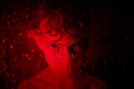 Photo for Through glass wall view of displeased child looking at camera in neon red light - Royalty Free Image