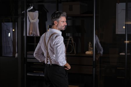 Téléchargez les photos : Homme aux cheveux gris d'âge moyen en chemise blanche classique et pantalon noir debout devant une porte en verre avec les mains à la taille et regardant ailleurs - en image libre de droit