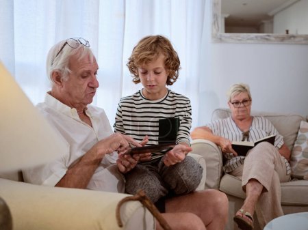 Téléchargez les photos : Focalisé grand-père et petit-fils naviguant tablette moderne tout en étant assis sur un canapé confortable dans le salon léger avec femme à la maison - en image libre de droit