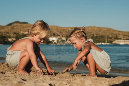 Téléchargez les photos : Vue de côté corps entier de focalisé pieds nus frère et sœur en vêtements d'été assis sur des hanches près du lac ondulant et creusant du sable par une journée ensoleillée - en image libre de droit